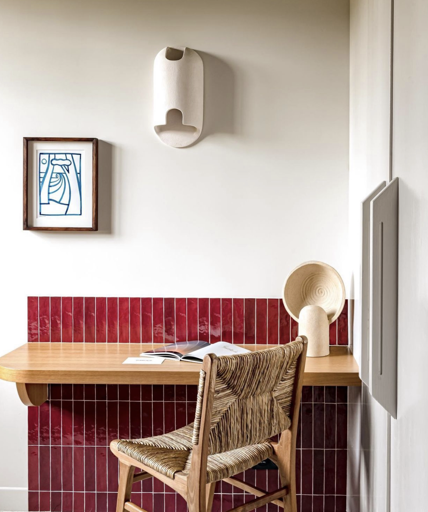 A desk with red tiled backsplash, a rattan chair, and decorative wall art including a framed drawing and sculptural objects in a minimal, modern style.