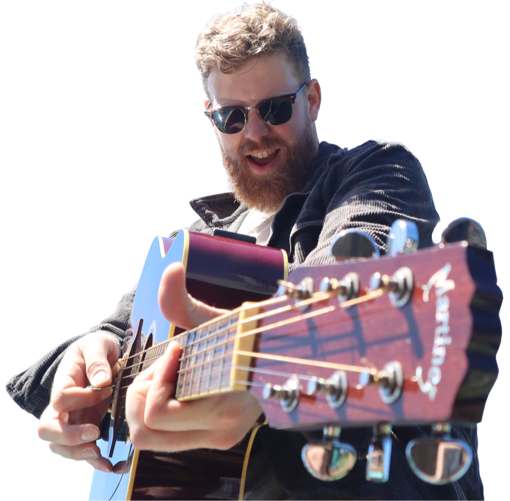 A man with sunglasses and a beard playing an acoustic guitar outdoors, sounding joyful.