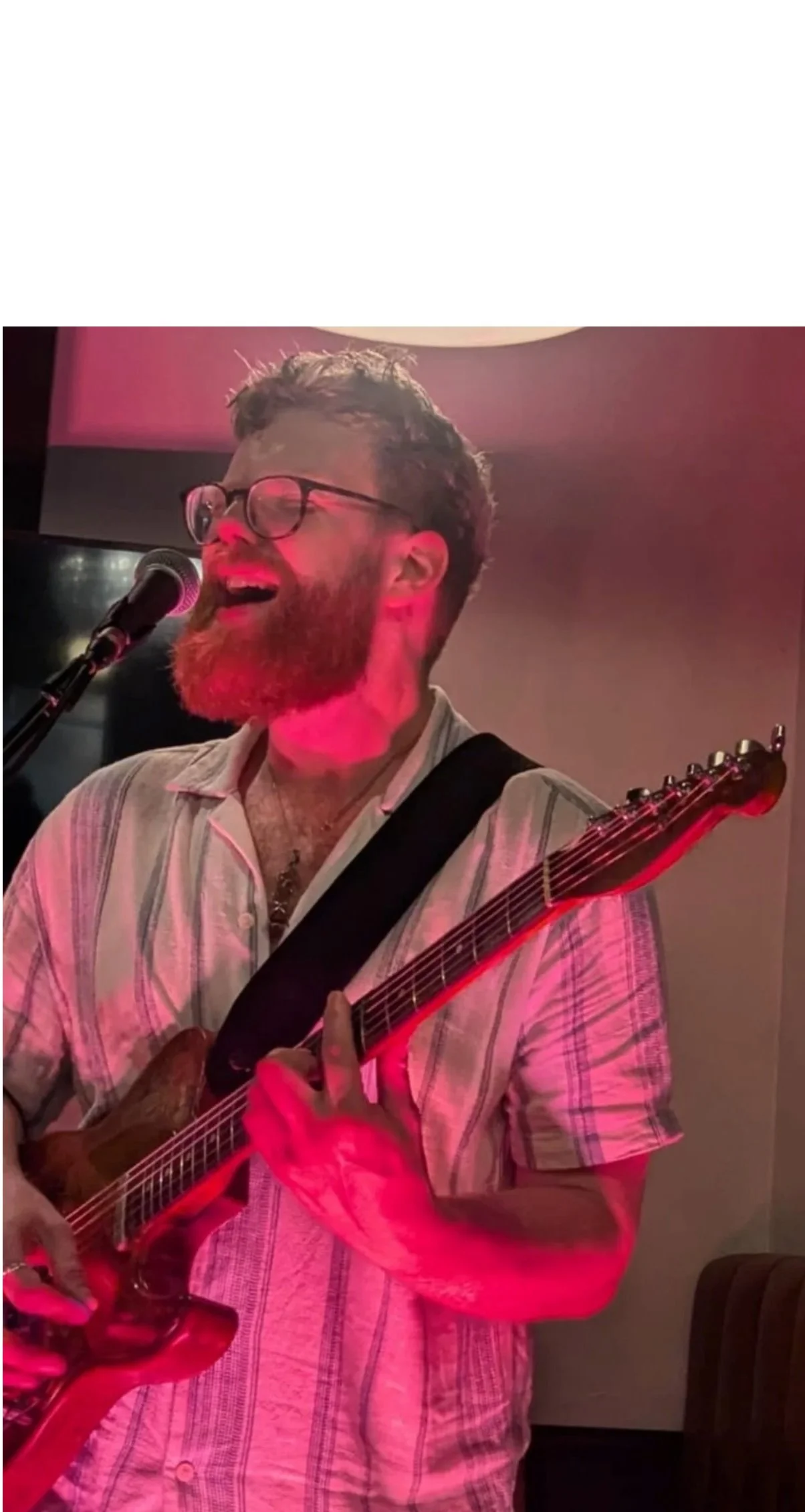 A man with glasses and a beard singing into a microphone while playing an electric guitar in a dimly lit room.