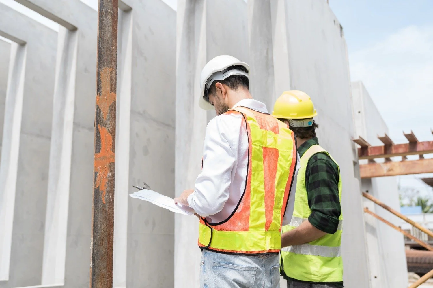 Two construction workers wearing safety helmets and high-visibility vests reviewing plans at a building site.