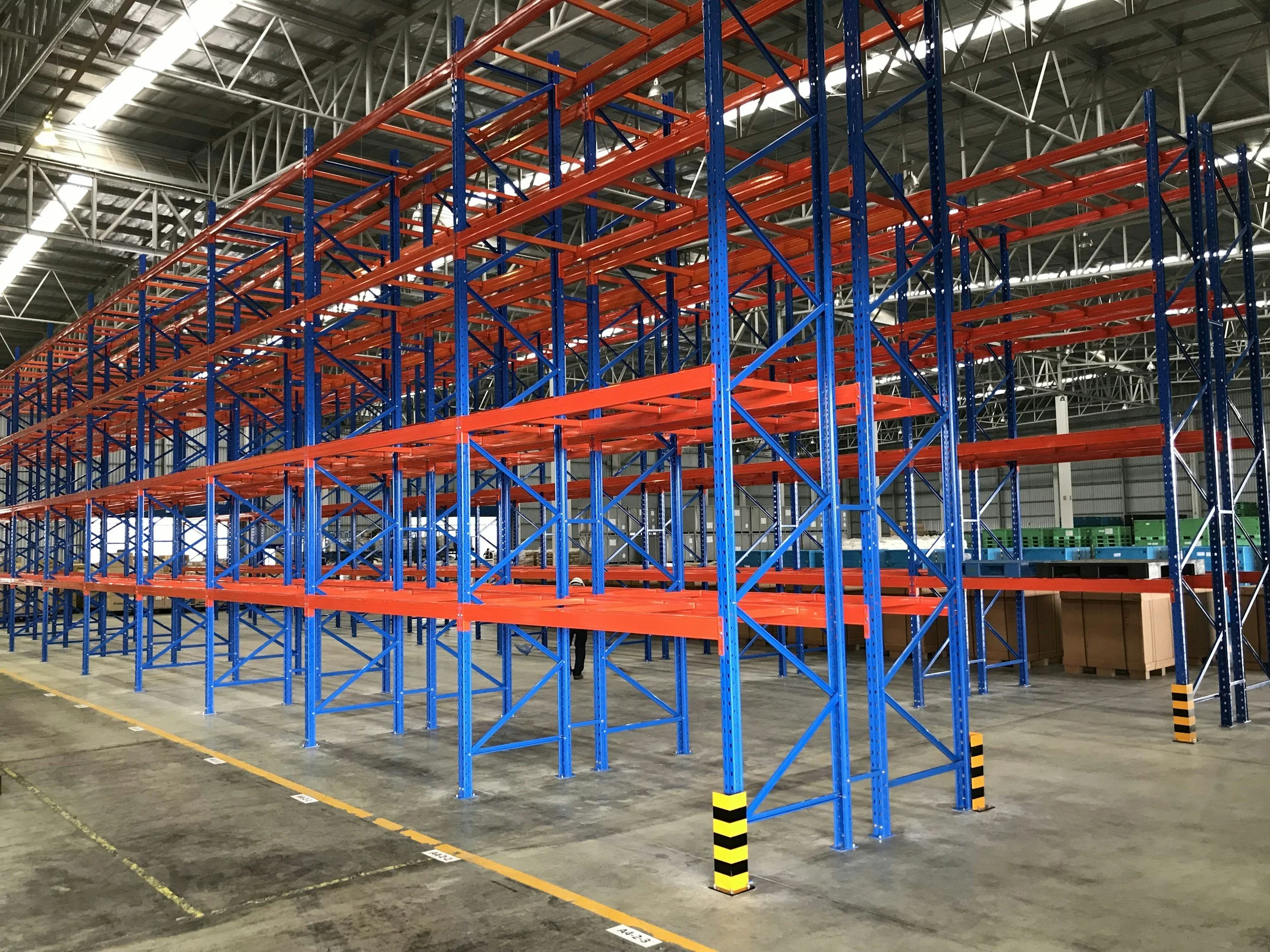 Interior of a warehouse with multiple levels of orange and blue metal shelving racks for storing goods, some boxes are visible at the bottom. The concrete floor has marked lines and labels, and the ceiling is made of metal beams with bright lighting.