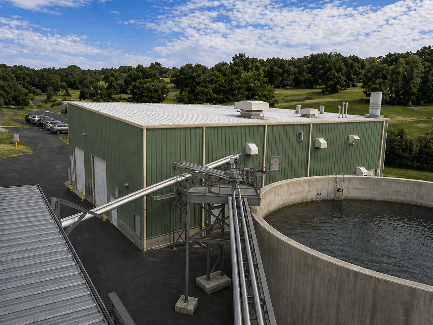 Industrial water treatment facility building with green walls and large circular concrete tank filled with water, surrounded by piping and equipment, with a parking lot and wooded landscape in the background.