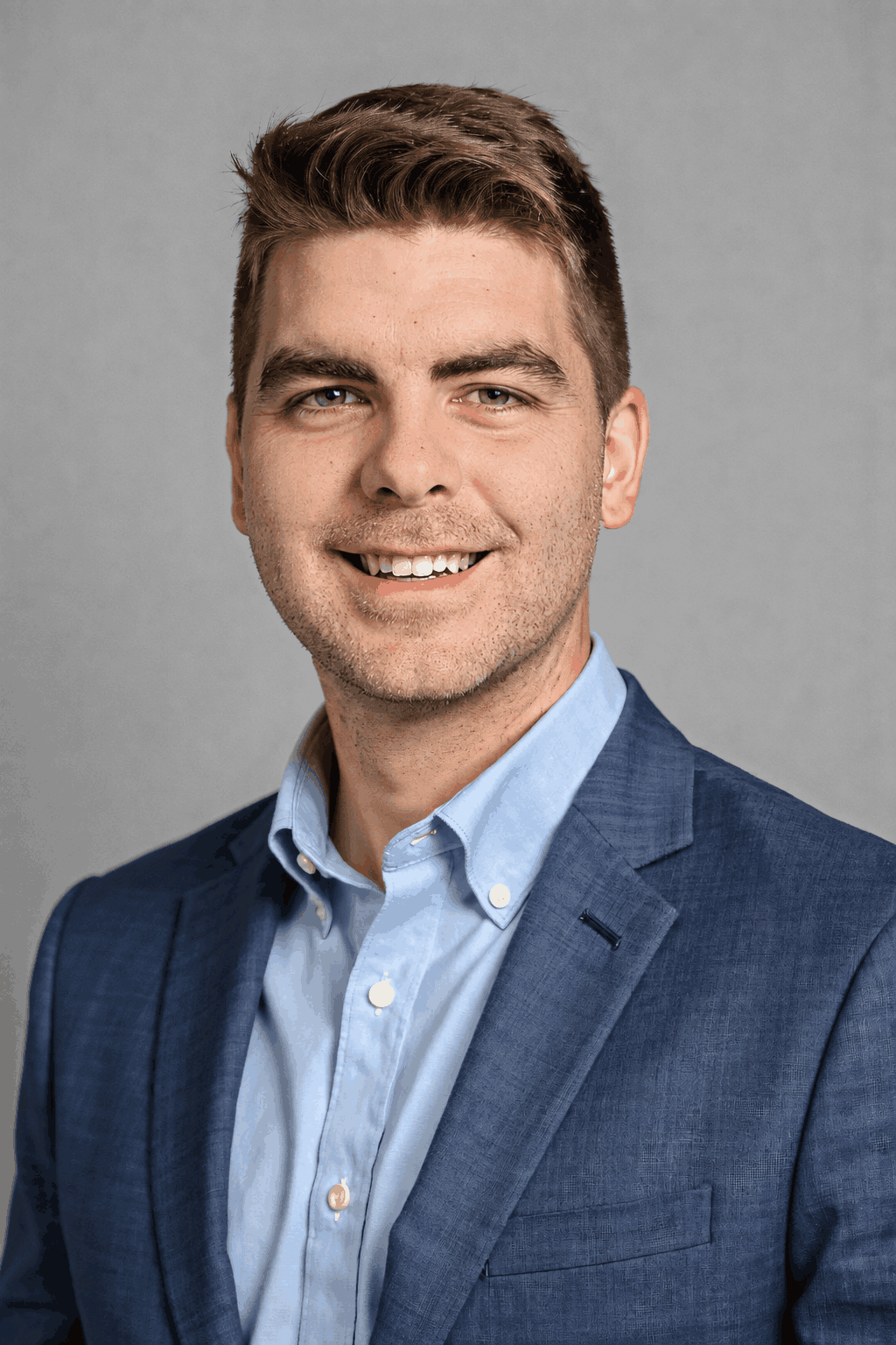 Portrait of Tyler Jewell, a young man with brown hair and blue eyes, smiling, wearing a blue suit and light blue shirt, against a gray background.