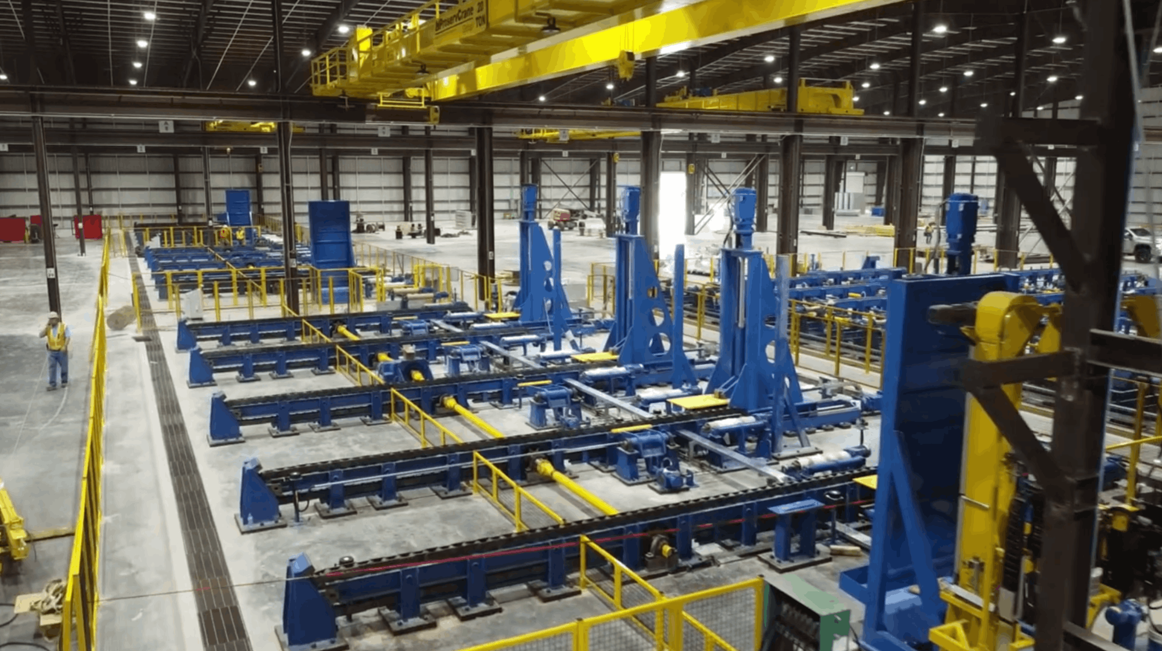 Large industrial warehouse with blue machinery, yellow safety railing, and overhead yellow cranes, along with a few workers in safety vests.