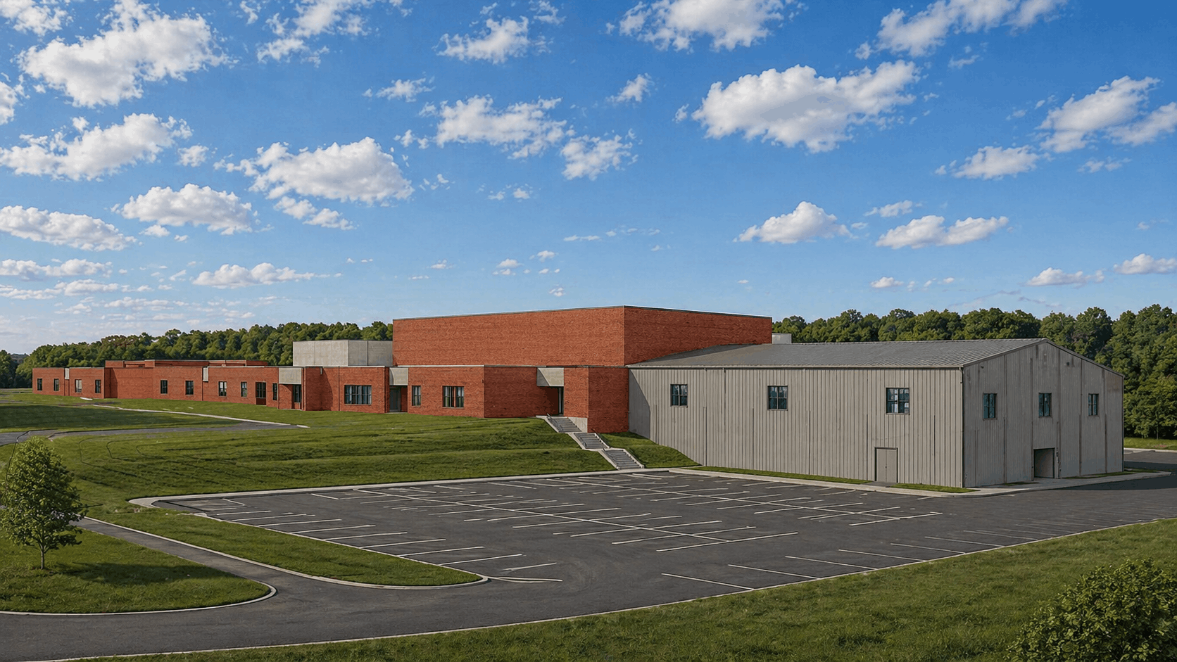 An empty parking lot in front of a modern school building with red brick and gray siding, surrounded by green grass and trees under a blue sky with scattered white clouds.