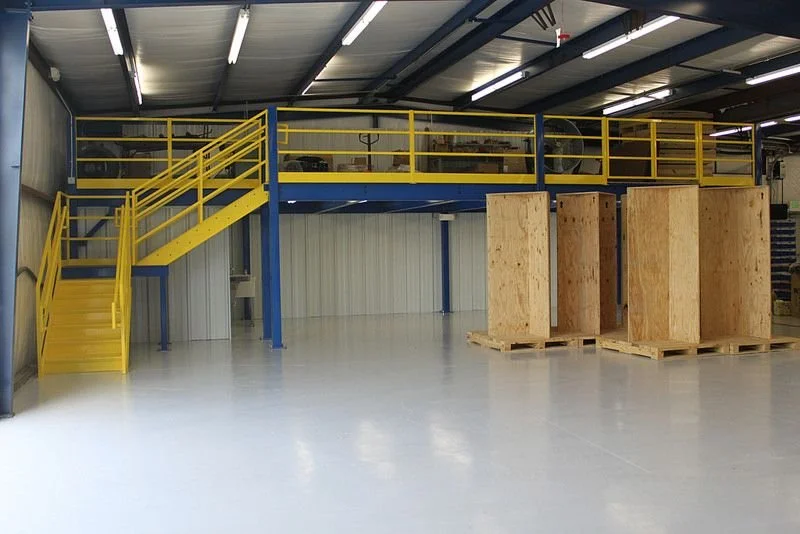 Industrial warehouse interior with yellow metal staircase and safety railings leading to a second-floor platform, alongside three large wooden crates on pallets.