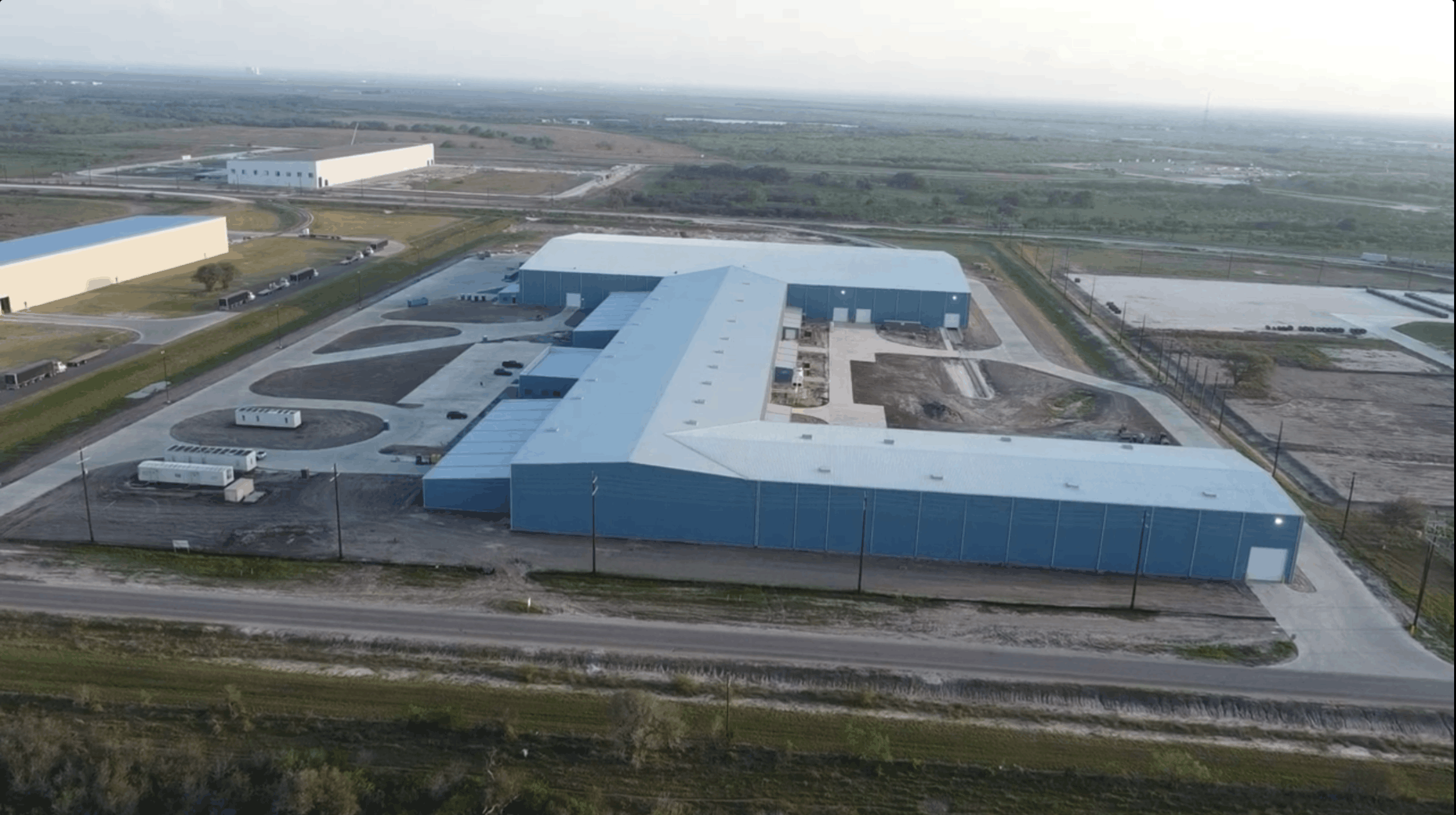 An aerial view of a large industrial or warehouse complex with multiple blue buildings, parking lots, and surrounding roads, extending into an open landscape with fields and sparse vegetation.