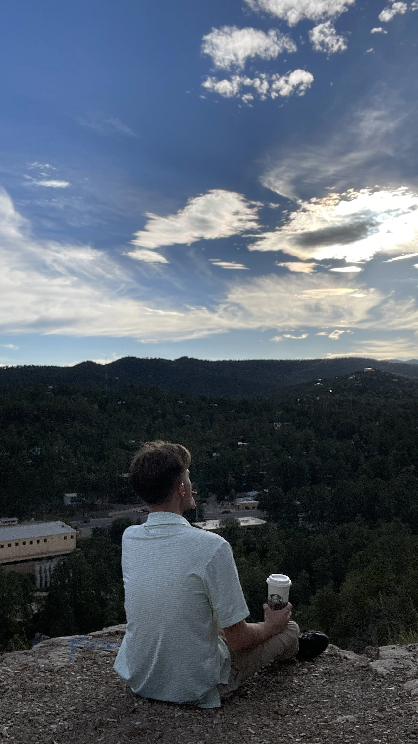 A person with short brown hair, sitting cross-legged on a rocky surface, holding a Starbucks coffee cup, overlooking a hilly landscape with trees and buildings under a partly cloudy sky during sunset or late afternoon.