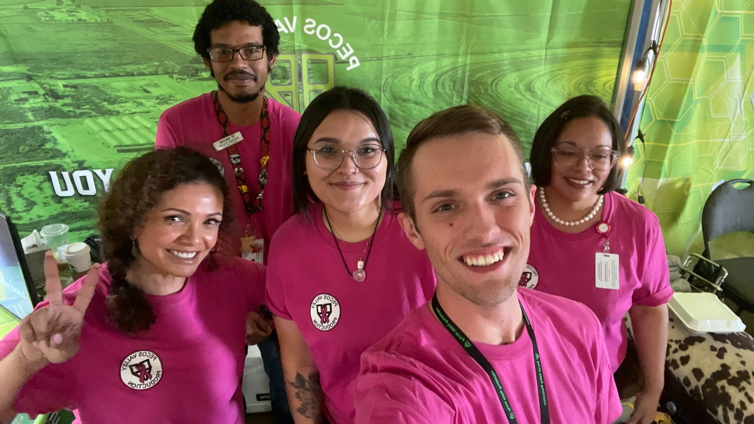 A group of five cannabis dispensary staff members wearing matching bright pink T‑shirts pose together in a booth with a green branded backdrop at an event, creating a friendly, professional team photo