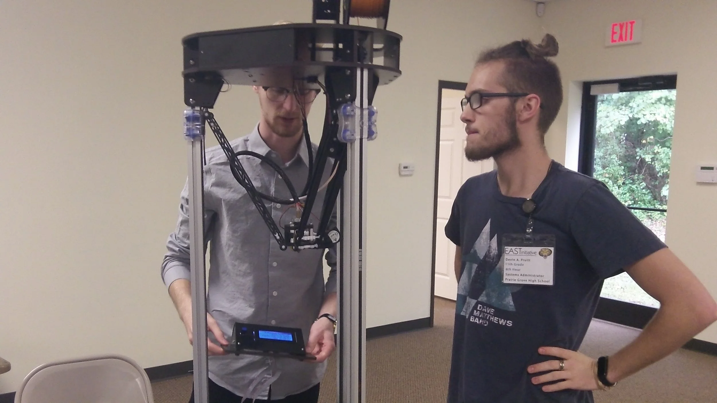 Two young men wearing glasses interact with a robotic device inside a room with beige walls and a window showing trees outside.