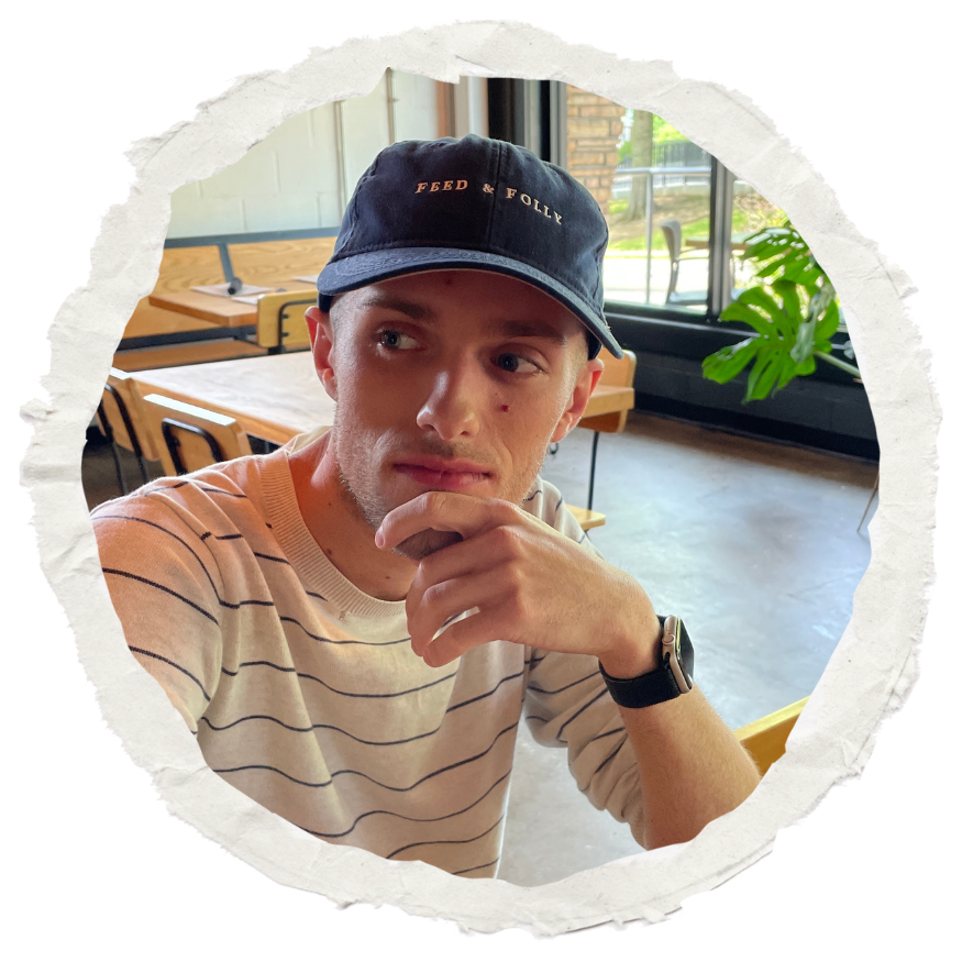 A man in a casual setting wearing a navy 'Feed & Folly' baseball cap and a tan striped t-shirt, posing indoors with his hand to his face in a thoughtful gesture. Modern wooden furniture and large windows with greenery visible in the background.