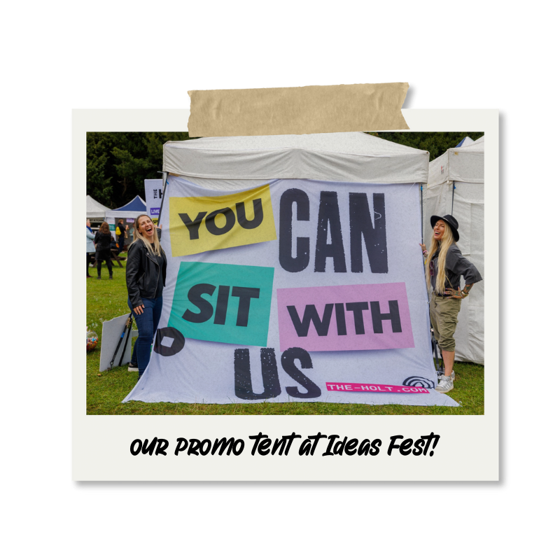 Two women standing in front of a promotional tent at Ideas Fest, with a sign that reads 'You Can Sit With Us,' in colorful block letters. One woman is smiling and wearing a leather jacket, while the other is laughing and wearing a hat and khaki pants.