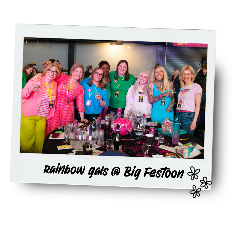 Group of women smiling and posing around a table at a big celebration event. They are wearing colorful outfits and lanyards. The table has drinks and decorations. Caption reads "rainbow gals @ Big Festoon" with flower icons.
