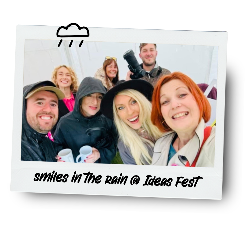 Group of seven people smiling and taking a selfie during a rainy outdoor event, some holding coffee mugs, with a caption "smiles in the rain @ Ideas Fest" at the bottom of the photo.