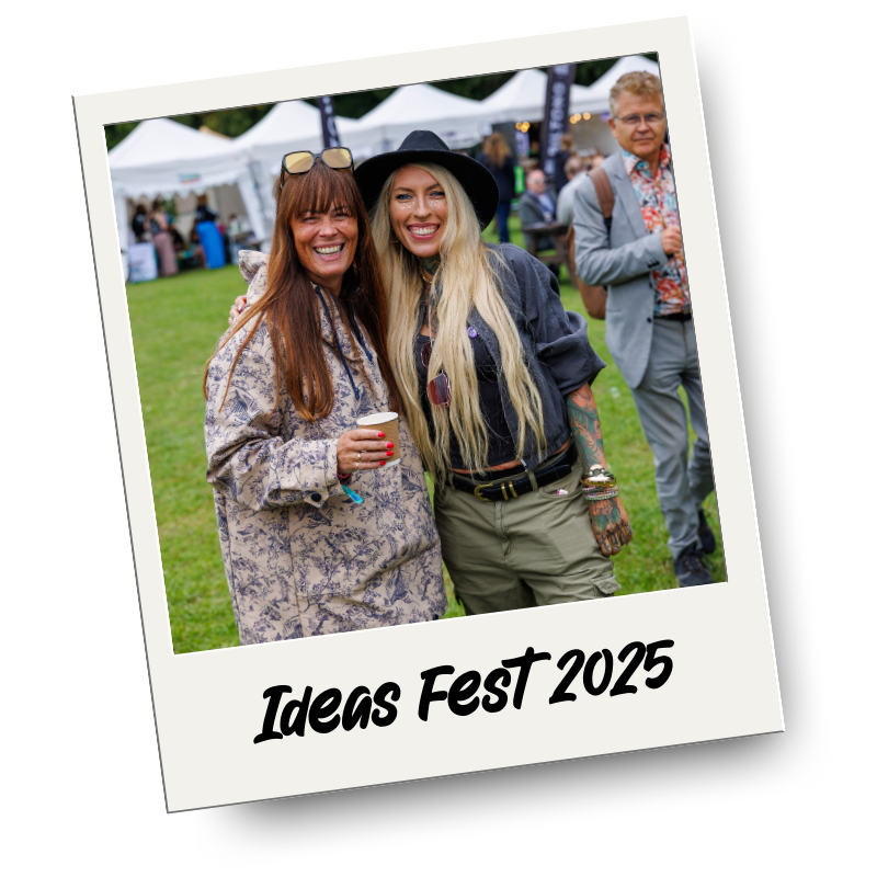 Two women smiling at Ideas Fest 2025 outdoors, with tents in the background. One woman has long brown hair and glasses on her head, the other has long blonde hair and a black hat. They appear to be enjoying the event.