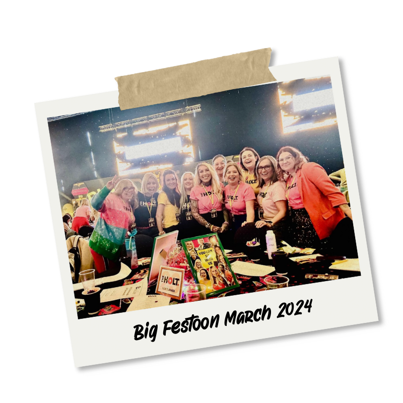 A group of women smiling and posing at a conference or event, with a table in front of them displaying various items and colorful posters. The background shows a stage with bright lights. The photo is framed as a polaroid with tape at the top and the caption "Big Festoon March 2024" written at the bottom.