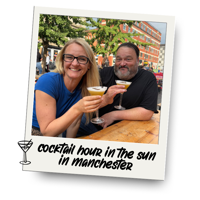 A smiling woman and man sitting outside at a wooden table, holding cocktail glasses, with trees and buildings in the background, enjoying a sunny day in Manchester.