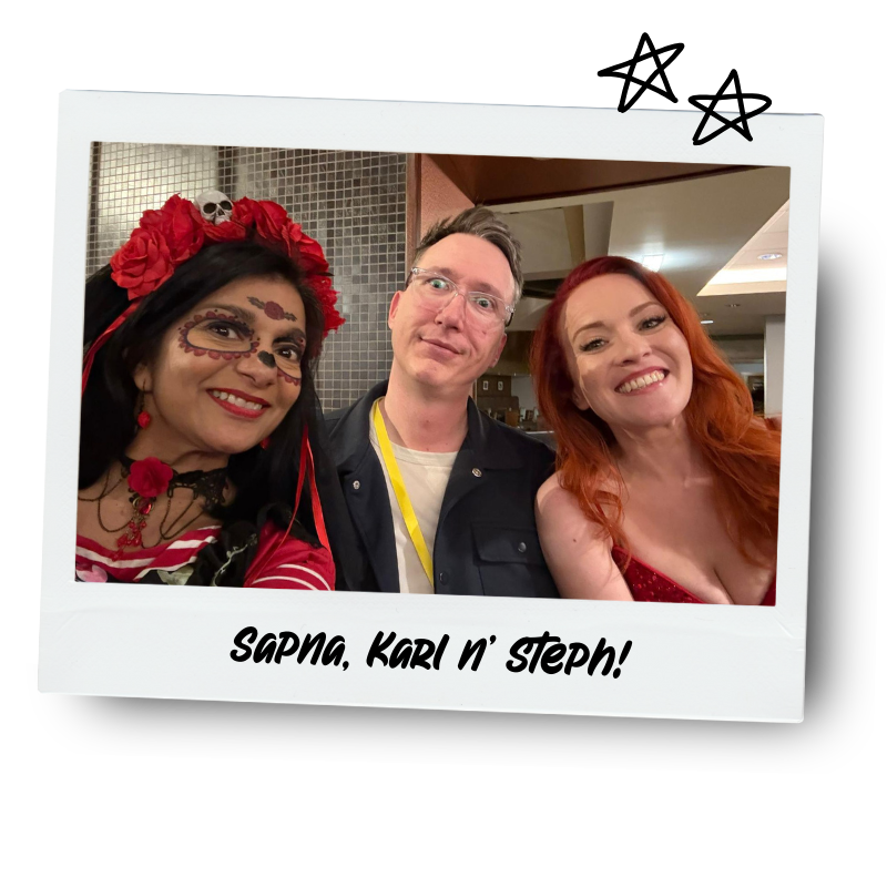 Three people smiling for a photo at a festive event. The woman on the left is dressed in a Day of the Dead-inspired costume with face paint, a red flower headpiece, and red accessories. The man in the middle is wearing glasses, a dark jacket, and a yellow lanyard. The woman on the right has long red hair and is wearing a red dress. The photo is framed with a white border and the caption "Sapna, Karl n' Steph!"