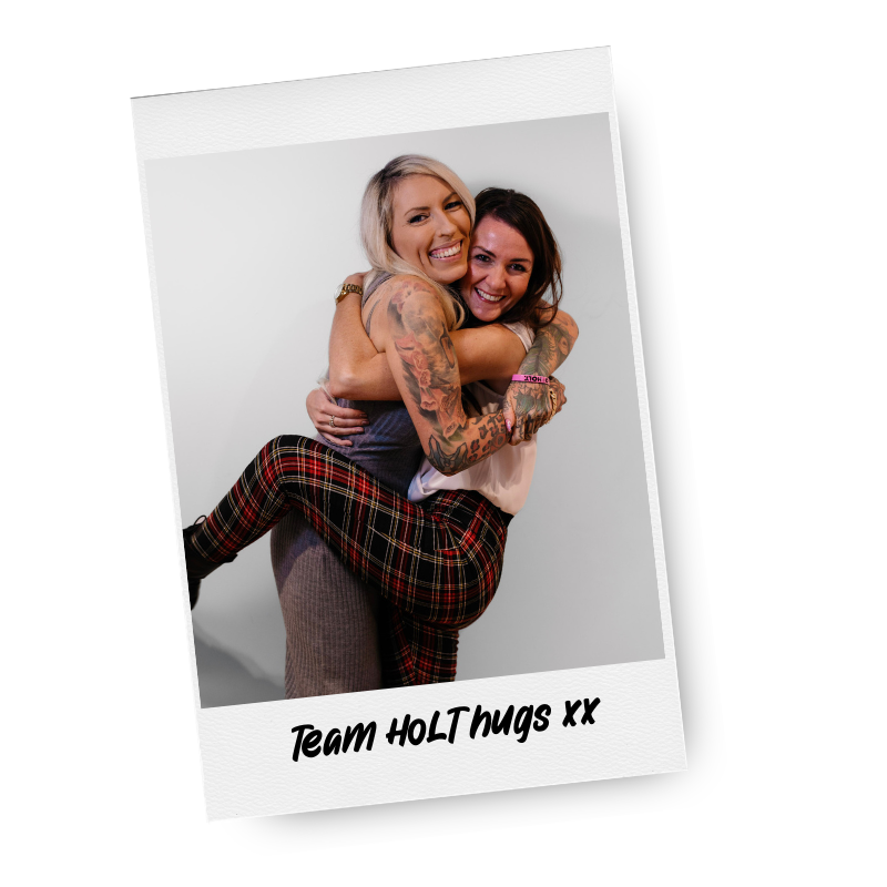 Two women embracing and smiling, one giving the other a piggyback ride, with tattoos on her arms, in front of a plain white background, with the caption "Team HOLThugs XX" at the bottom.