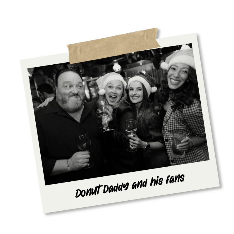 Group of four people celebrating with drinks, wearing Santa hats, at a holiday party, in black and white, with the caption 'Donut Daddy and his fans'.