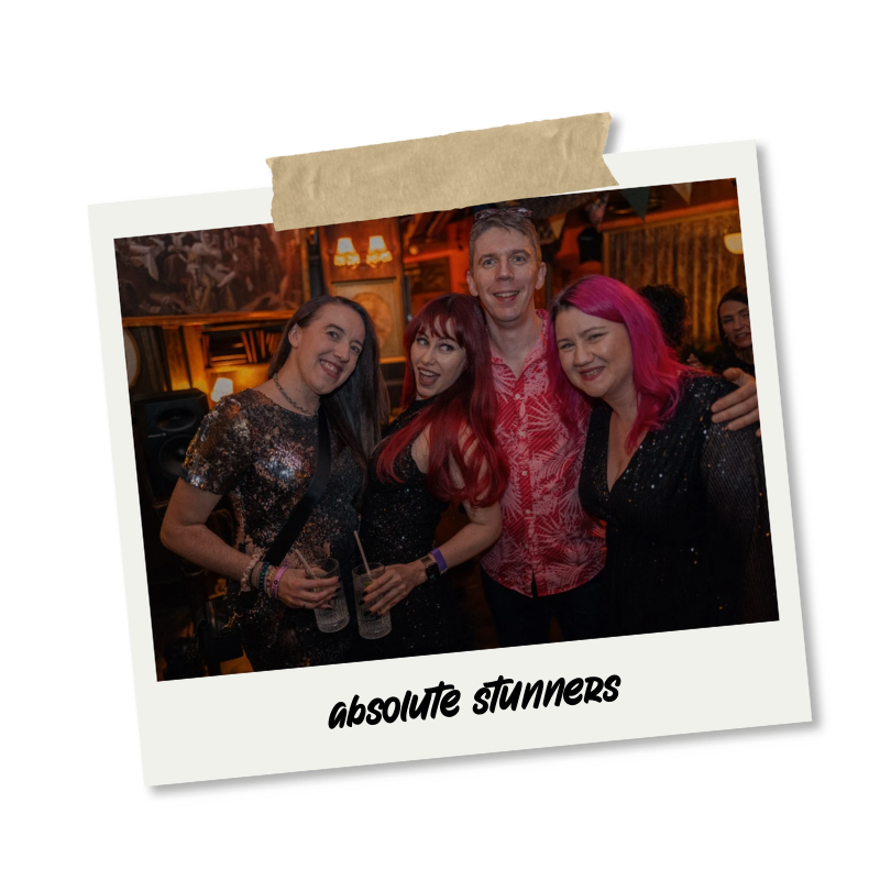Group of four friends celebrating at a party, smiling, holding drinks, in a cozy, warm-lit venue with wood decor, with the caption "absolute stunners"