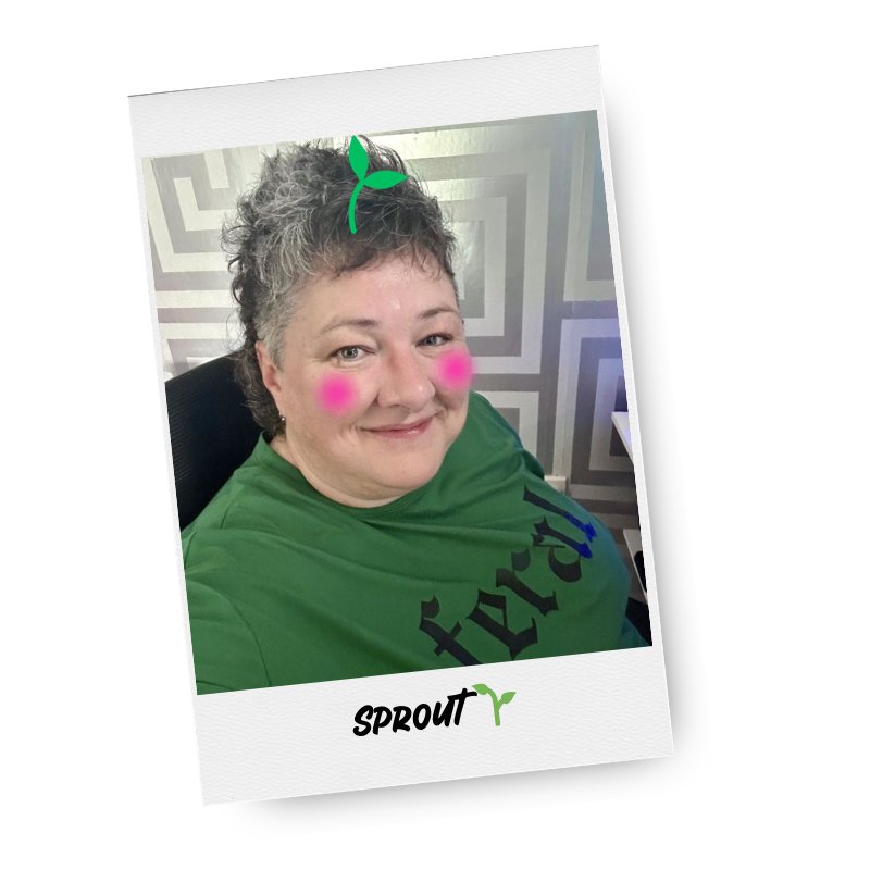 A woman with short, curly gray hair, wearing a green shirt with black text, sitting in a black chair, smiling. She has pink cheeks and a small green sprout graphic edited near her head.