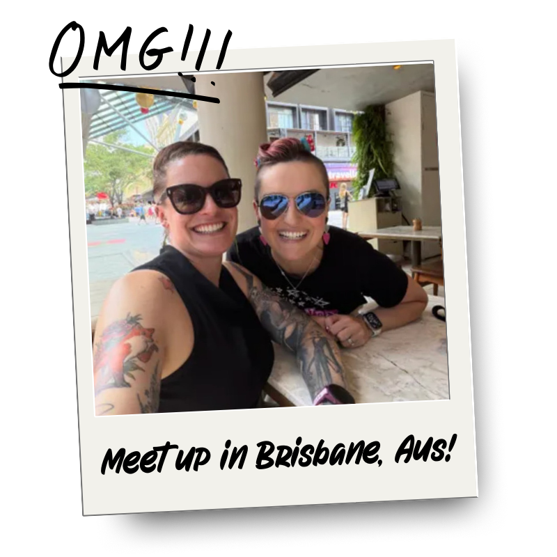 Two women smiling, wearing sunglasses, sitting at a table in a cafe or restaurant, with an outdoor scene visible through the window behind them. The photo has a Polaroid-style border with the caption "Meetup in Brisbane, Aus!"