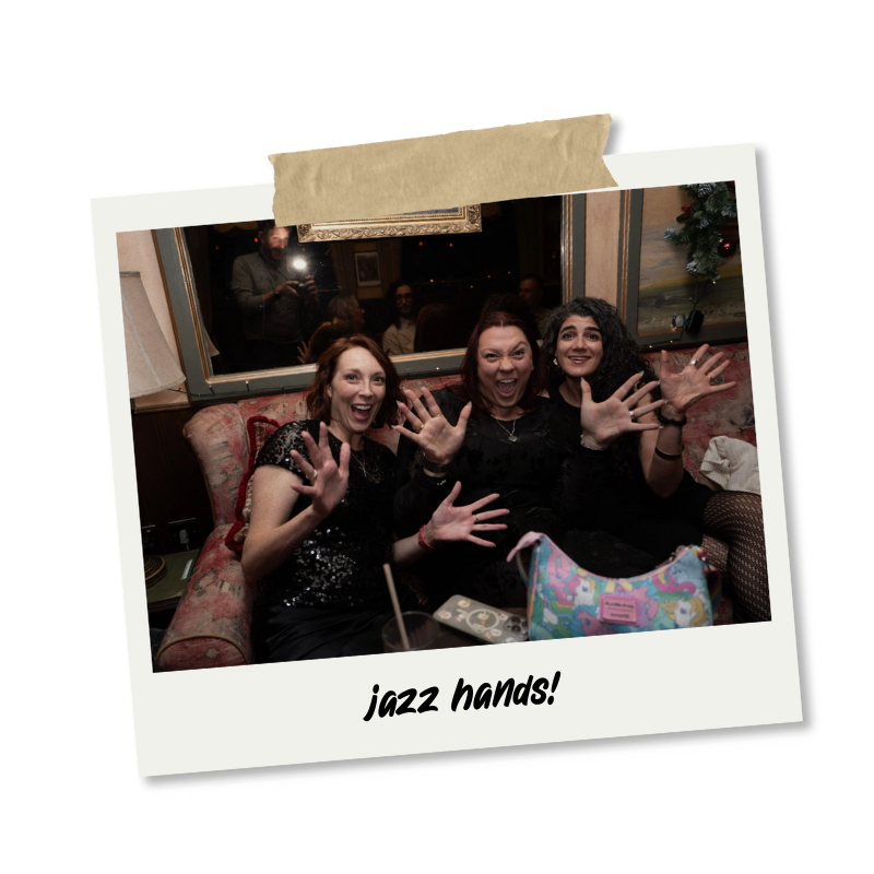 Three women sitting on a vintage sofa, smiling and waving their hands at the camera, at a social gathering. The photo has a Polaroid style border with the caption "jazz hands!" written at the bottom.