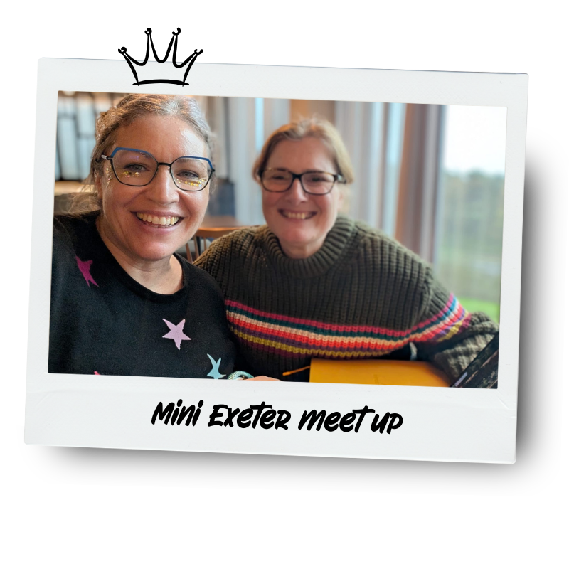 Two women smiling and taking a selfie at a mini Exeter meetup, sitting at a table with a window in the background.