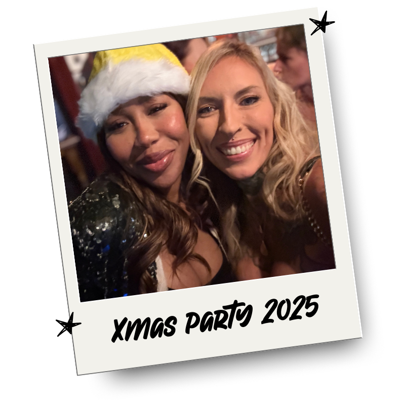 Two women smiling and taking a selfie at a Christmas party, one wearing a Santa hat, with a festive background.