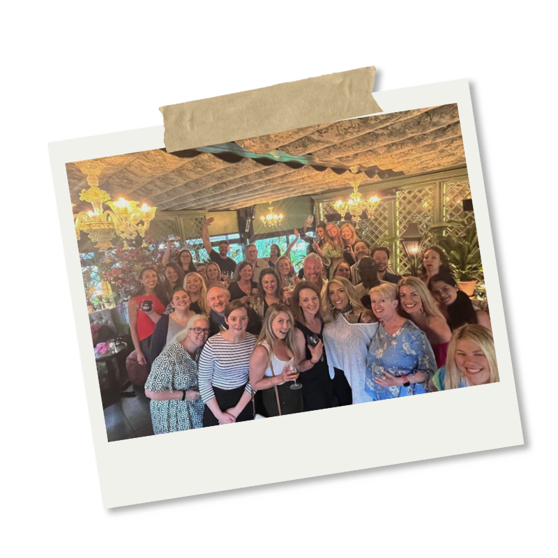 A large group of people gathered in a warmly lit restaurant or event space, posing for a group photo with smiling faces.