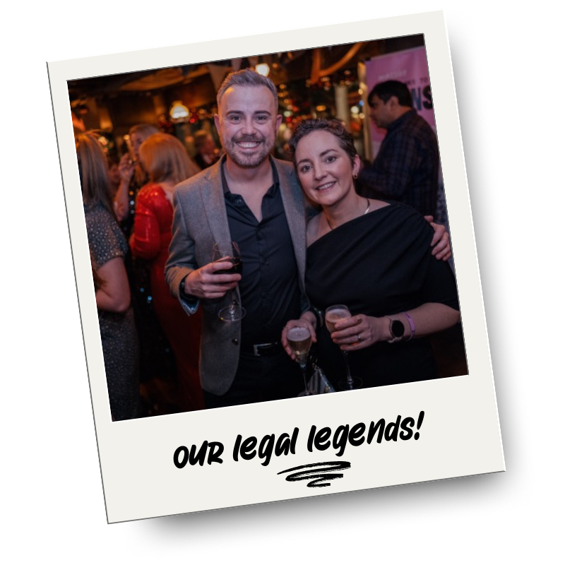 A man and woman smiling and holding glasses of wine at a social gathering or party, with other guests in the background. The photo has a caption below that reads 'Our legal legends!'.