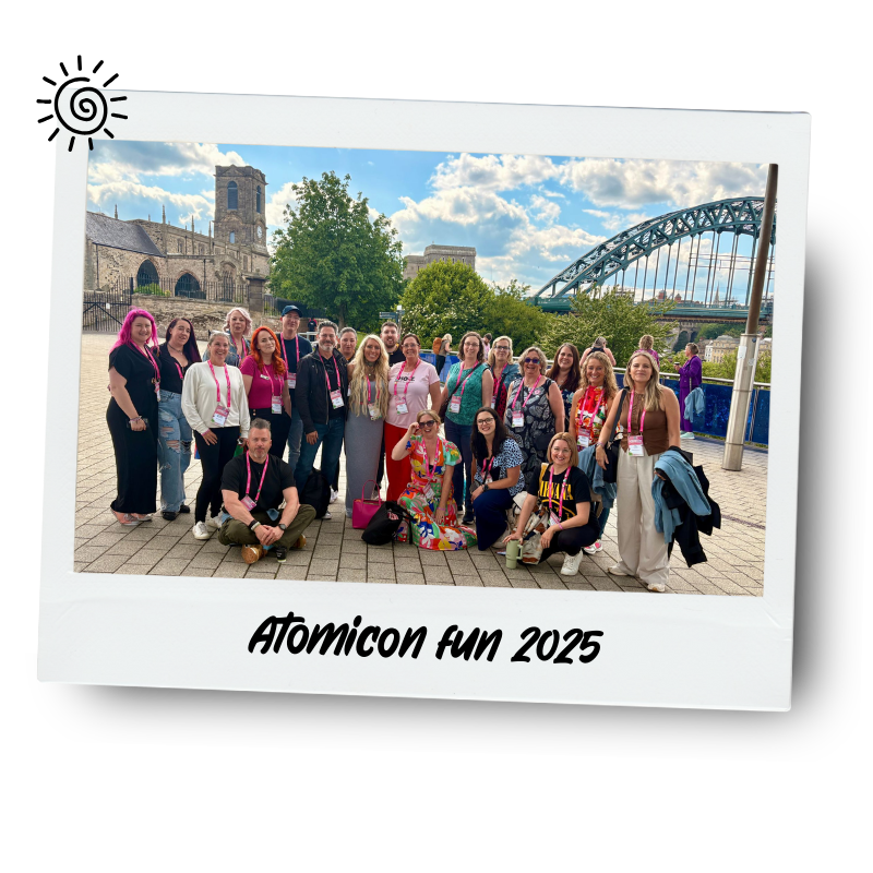 Group of people posing outdoors near a river with a bridge and historic building in the background, during daytime. The photo is framed like a polaroid with handwritten-style label "Atomicon fun 2025" at the bottom.
