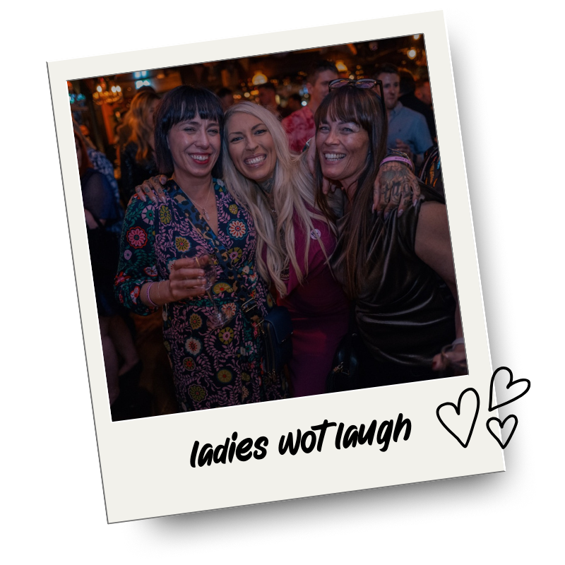 Three women smiling and laughing together at a social event or party, with a lively background and warm lighting.