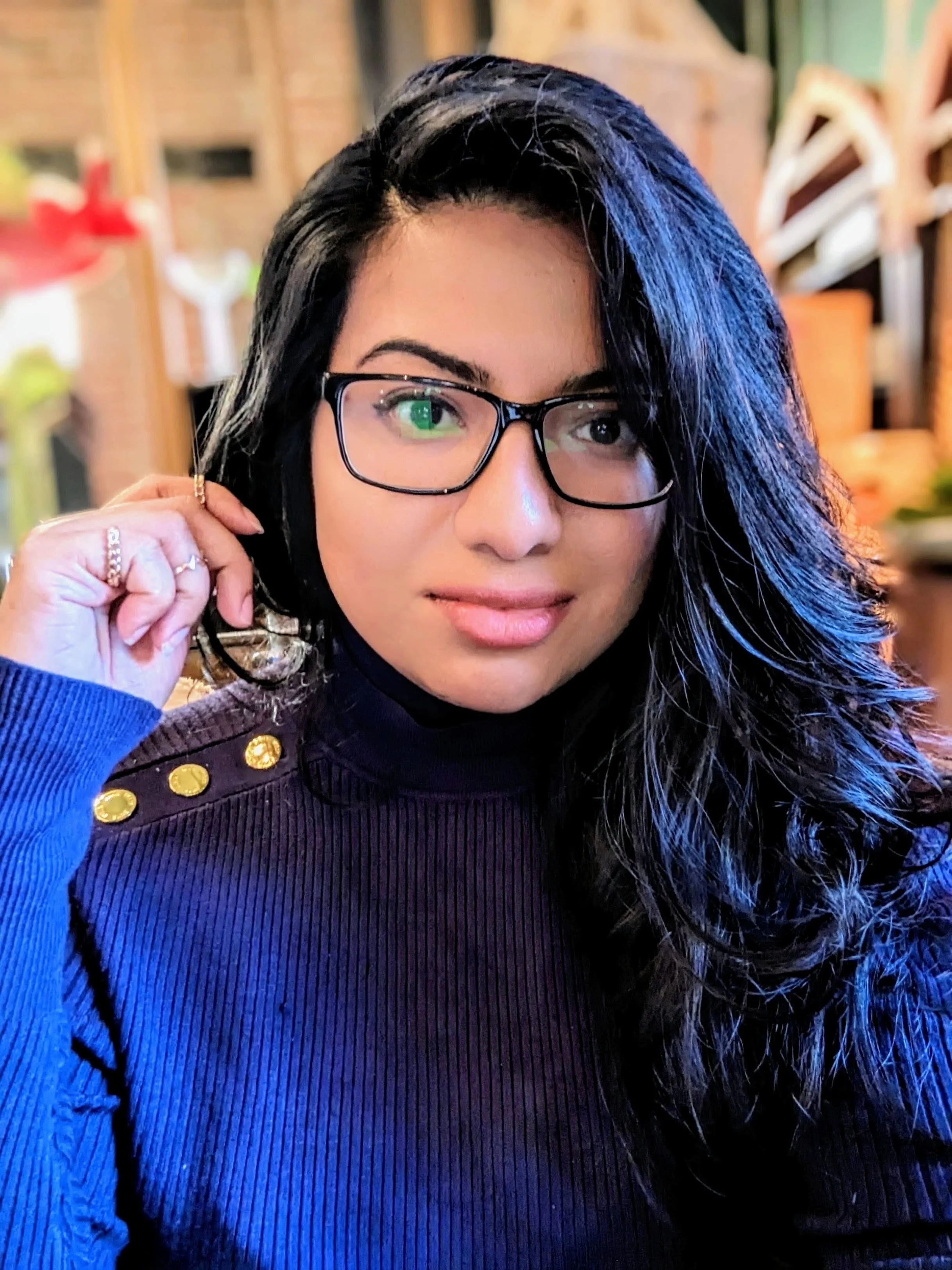 A woman with long black wavy hair, wearing black glasses and a blue turtleneck sweater with gold buttons on the shoulder, sitting indoors with a blurred background.