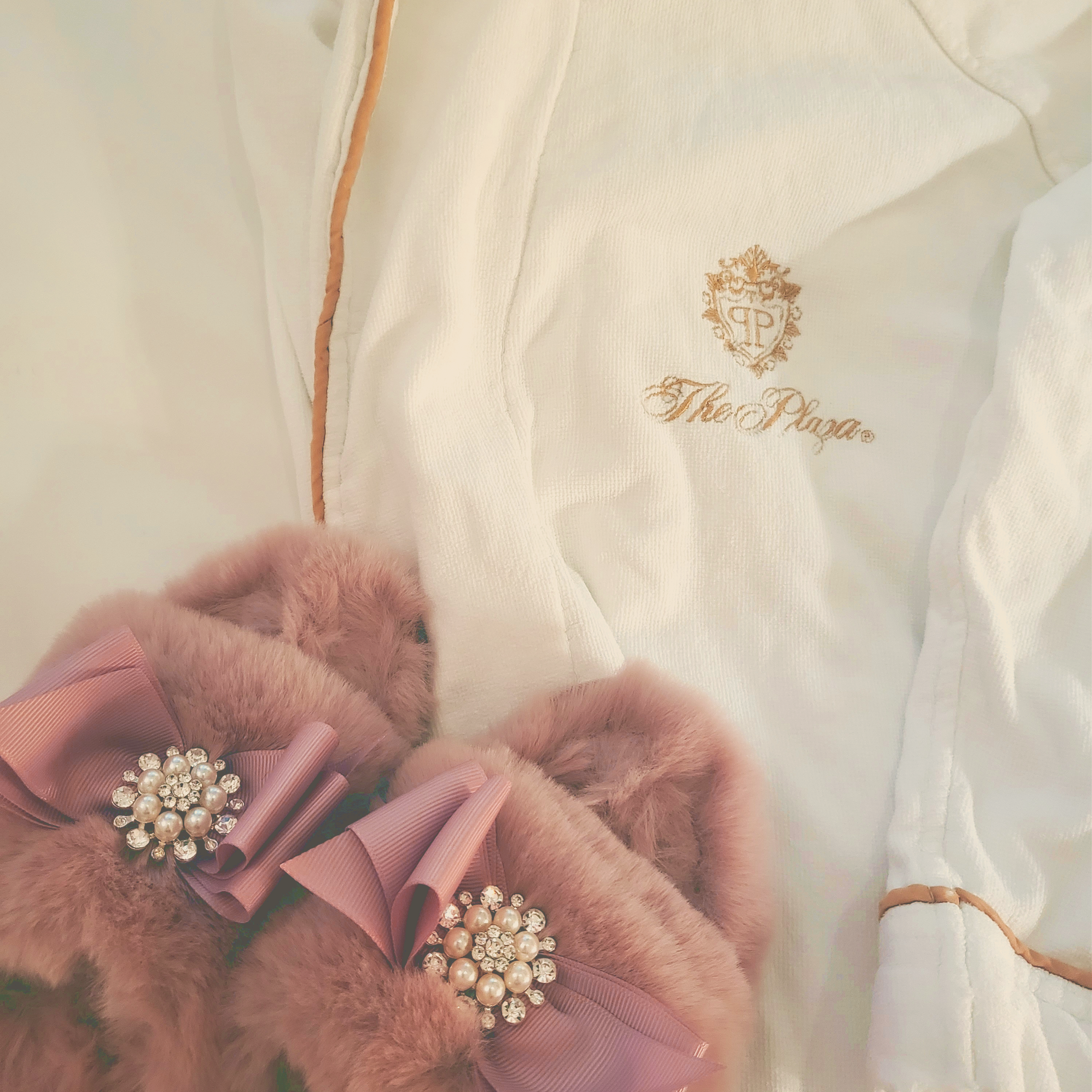 A closeup of a white robe with gold detail from The Plaza and a pair of fuzzy pink slippers with a matching grosgrain bow and pearl and diamond buttons
