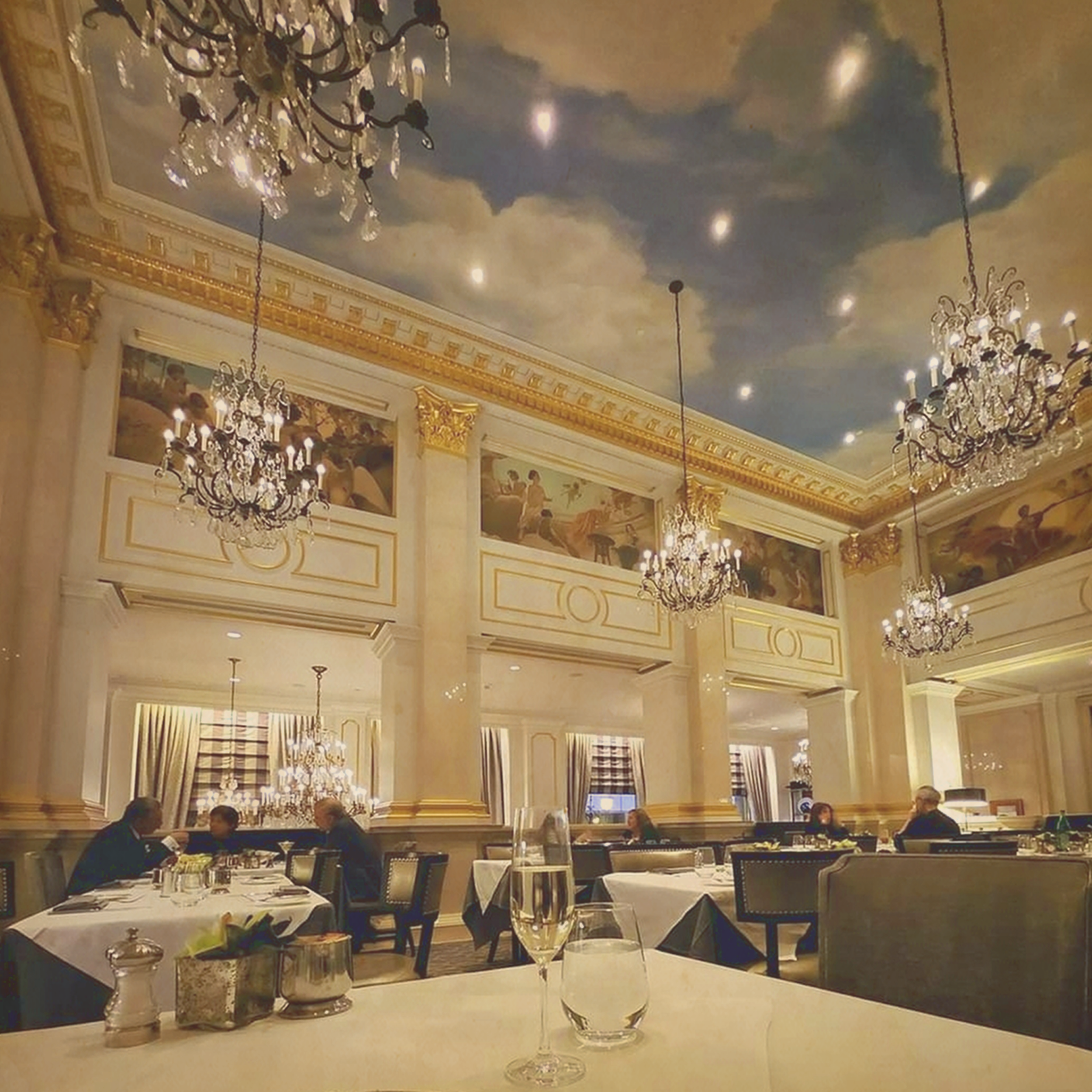 Astor Court in The St. Regis Hotel . A glass of champagne & a water on a white tablecloth in a Gilded Age courtyard style room with a cloud celling mural & Italianate frescos along the top. Gilt & marble columns &  5 chandeliers