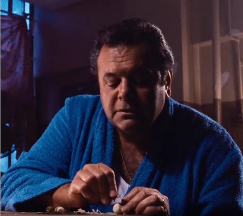 A man with dark, styled hair wearing a blue robe, sitting at a table in a dimly lit room, looking down with a serious expression.