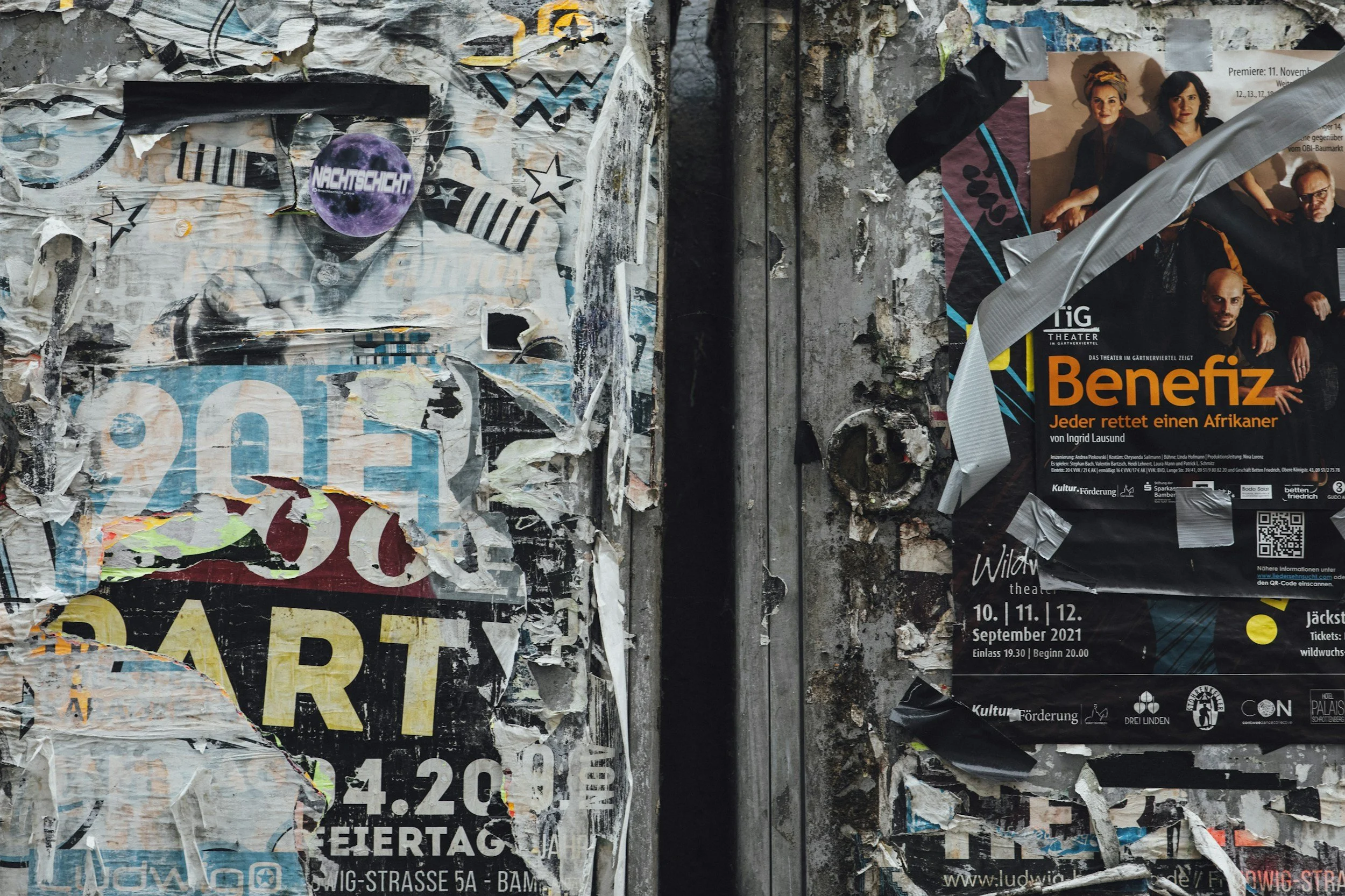 A wall covered in torn posters and flyers, with various colors and text, including an event poster for theatrical performances with the title "Benefiz" and images of performers.