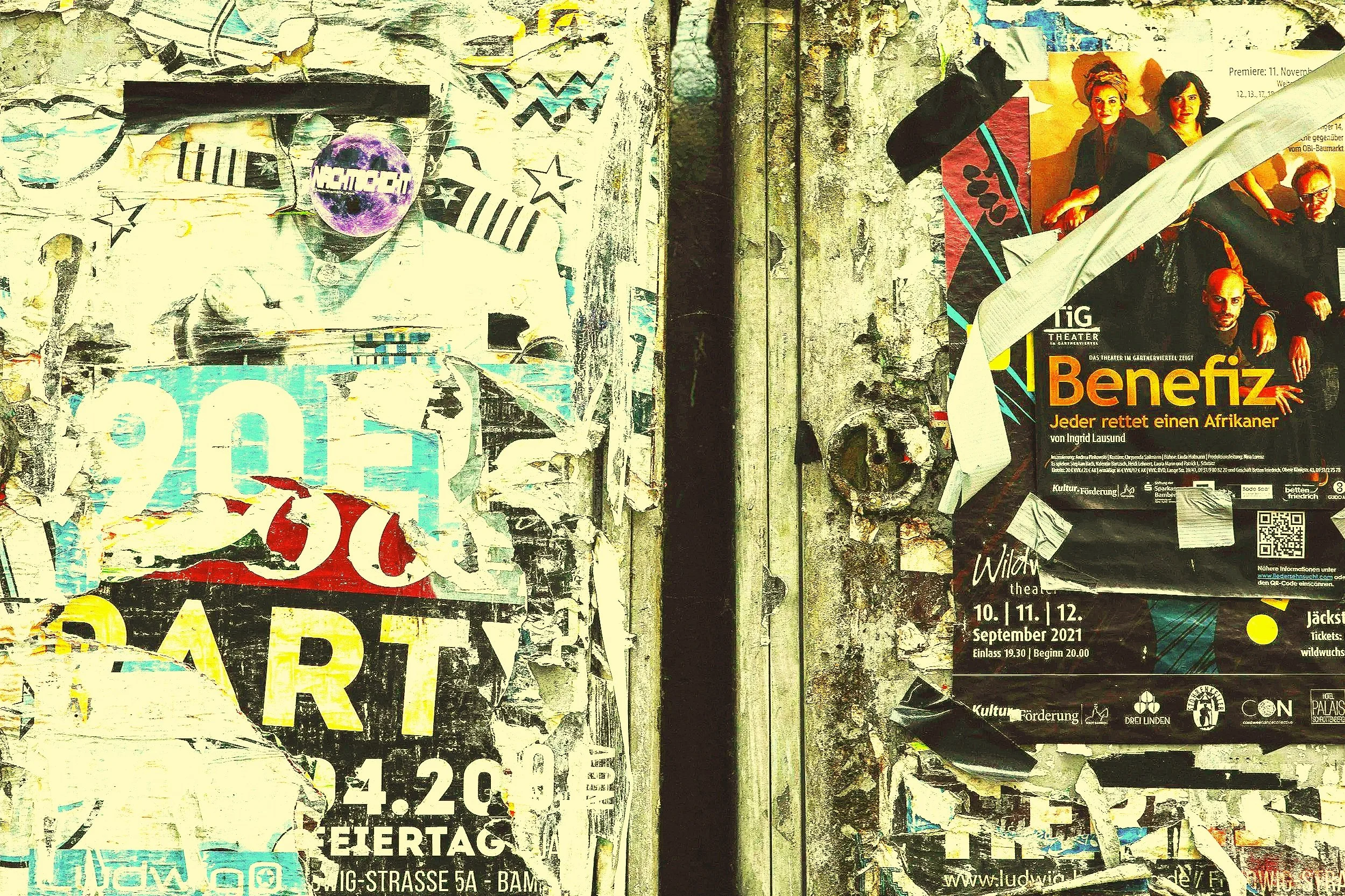 A wall covered with layered, torn posters and flyers, including a notice for a theatrical performance titled 'Benefiz' featuring a group of people, and various other partially visible advertisements and graffiti.
