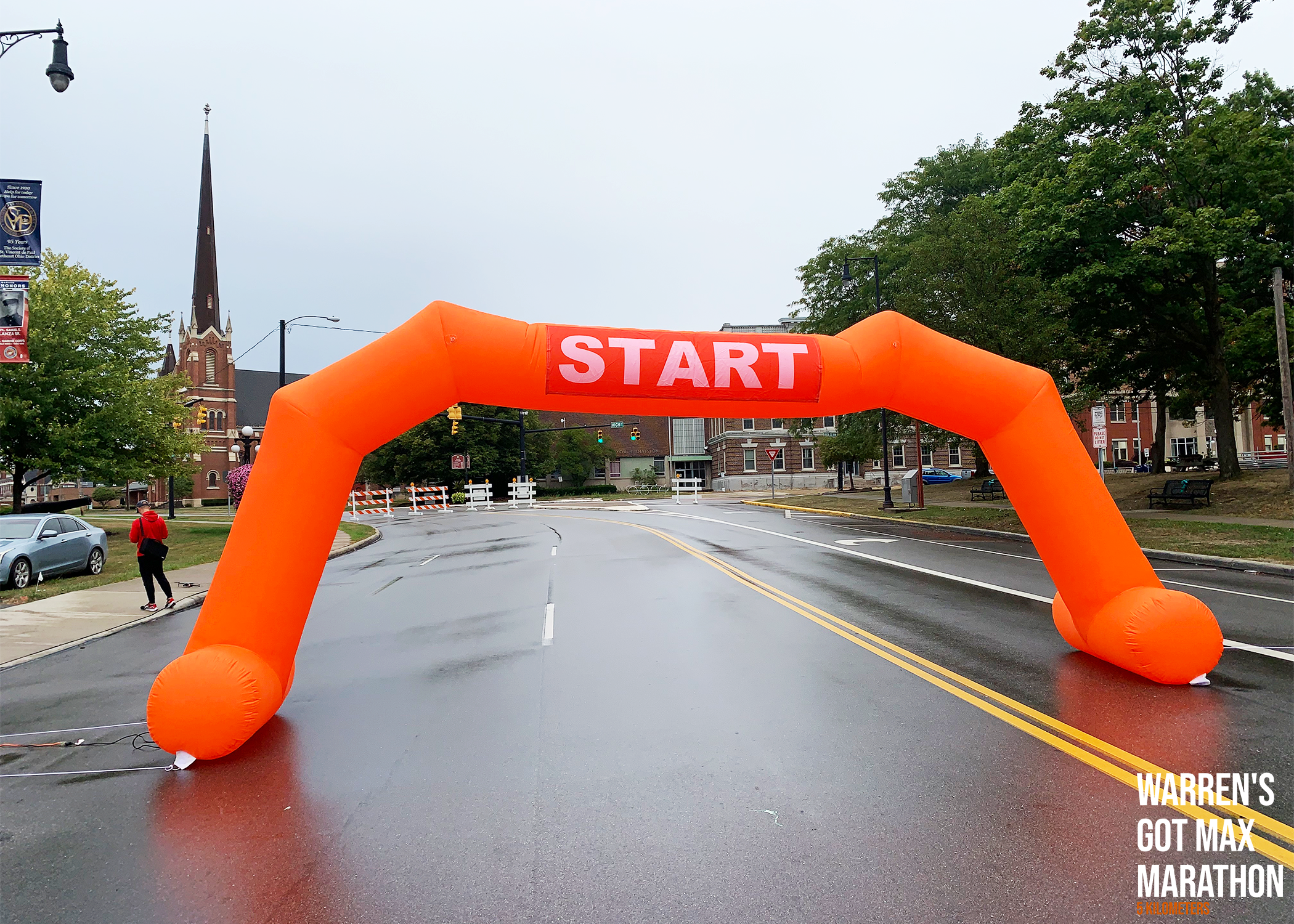 Orange inflatable start arch with the word "START" printed in white across the top, set up on a wet road in a town square with historic buildings, trees, and a church with a tall steeple in the background.
