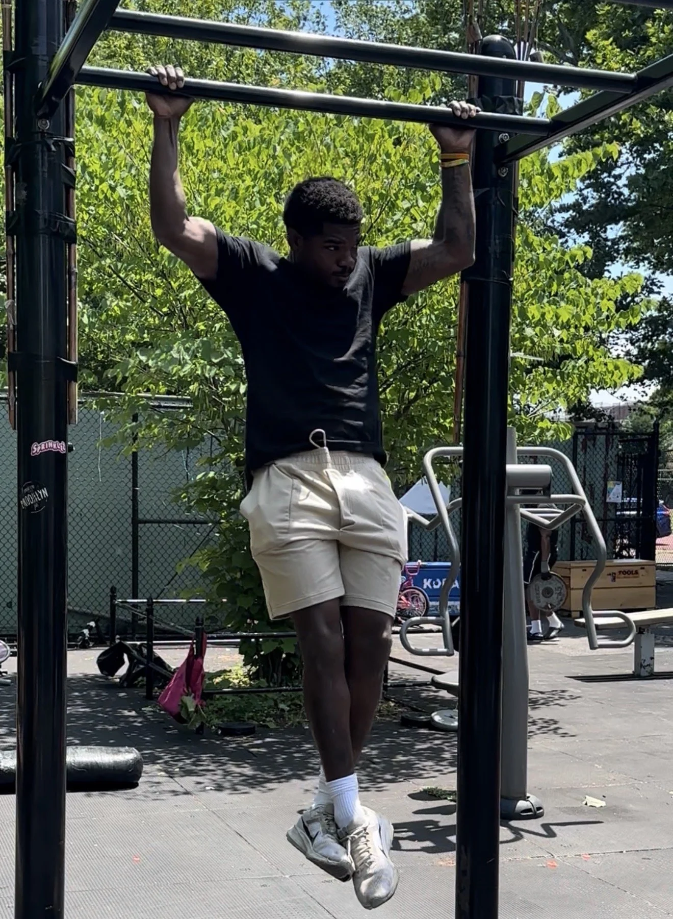Young man in black t-shirt and beige shorts doing pull-ups on outdoor gym equipment in a park with green trees and a chain-link fence in the background.