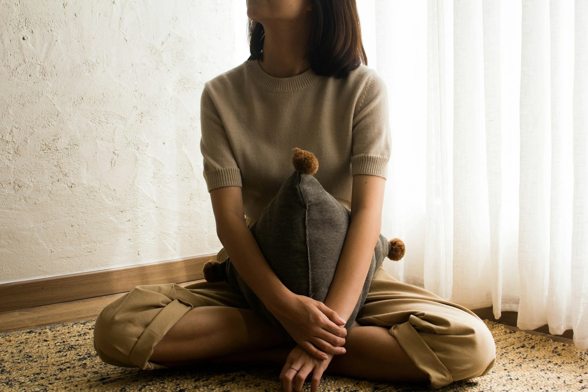 A woman sitting cross-legged on a carpeted floor, holding a gray pillow with brown pom-poms, near a cream-colored wall and sheer curtains.
