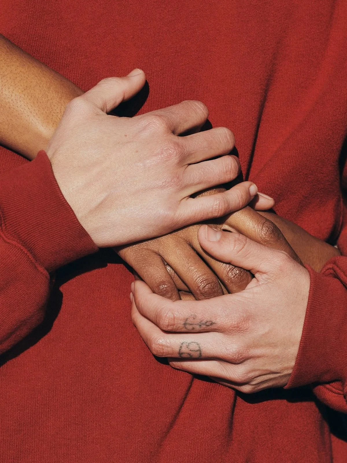 Close-up of three hands of different skin tones layered over a red fabric background, with one hand having a small tattoo on the fingers.