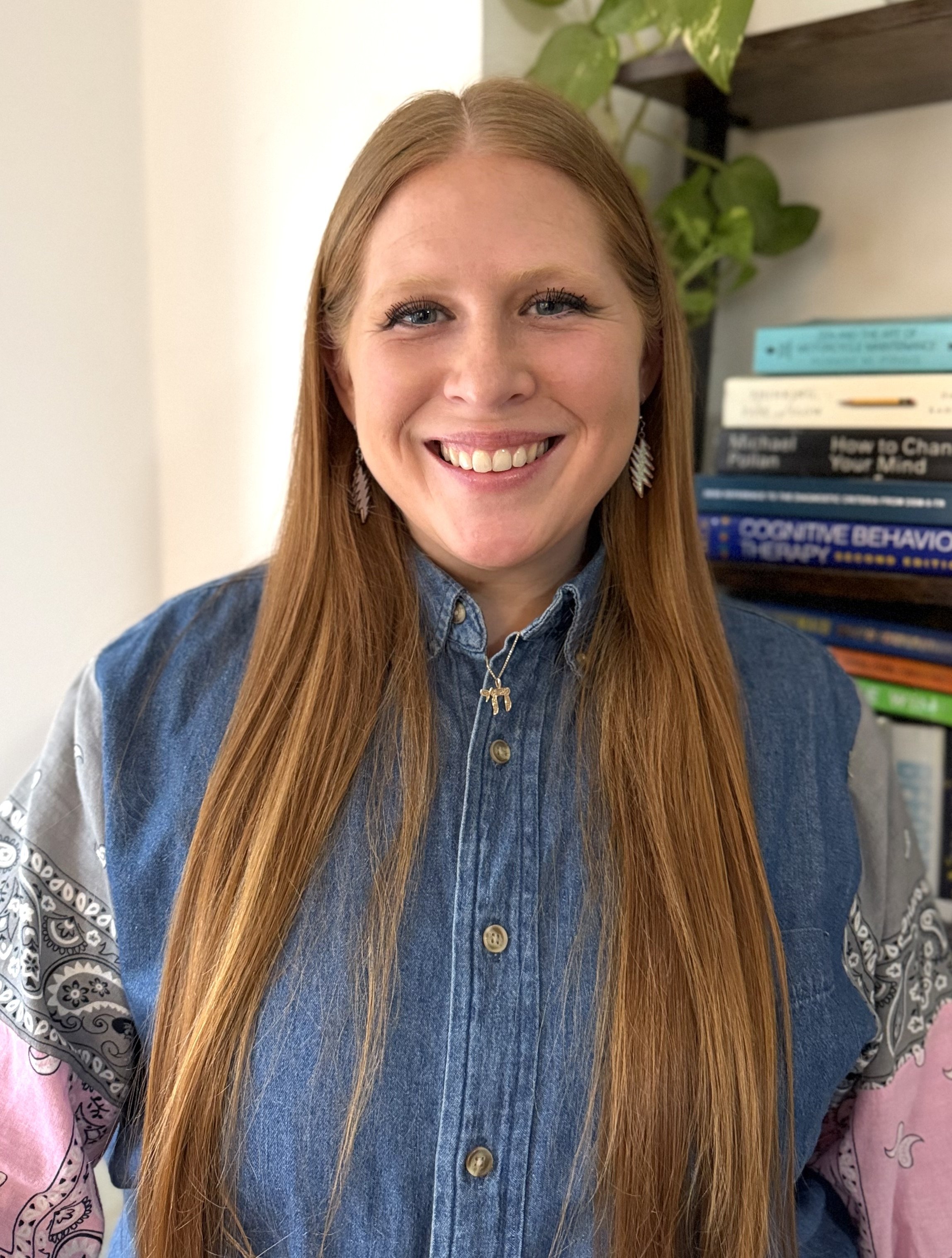 A woman with long red hair wearing a denim shirt with patches on the sleeves and a necklace with a dog pendant, smiling in front of a bookshelf.