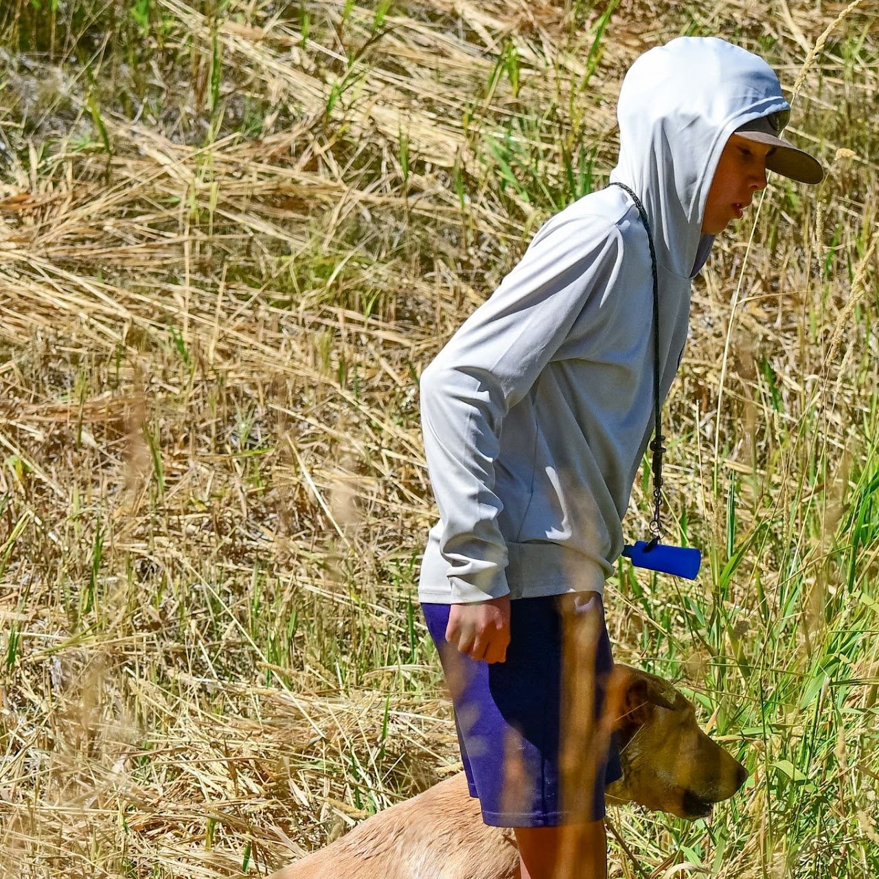 A person wearing a white hoodie, dark shorts, and a cap, standing in a grassy, dried field with a dog at their side.