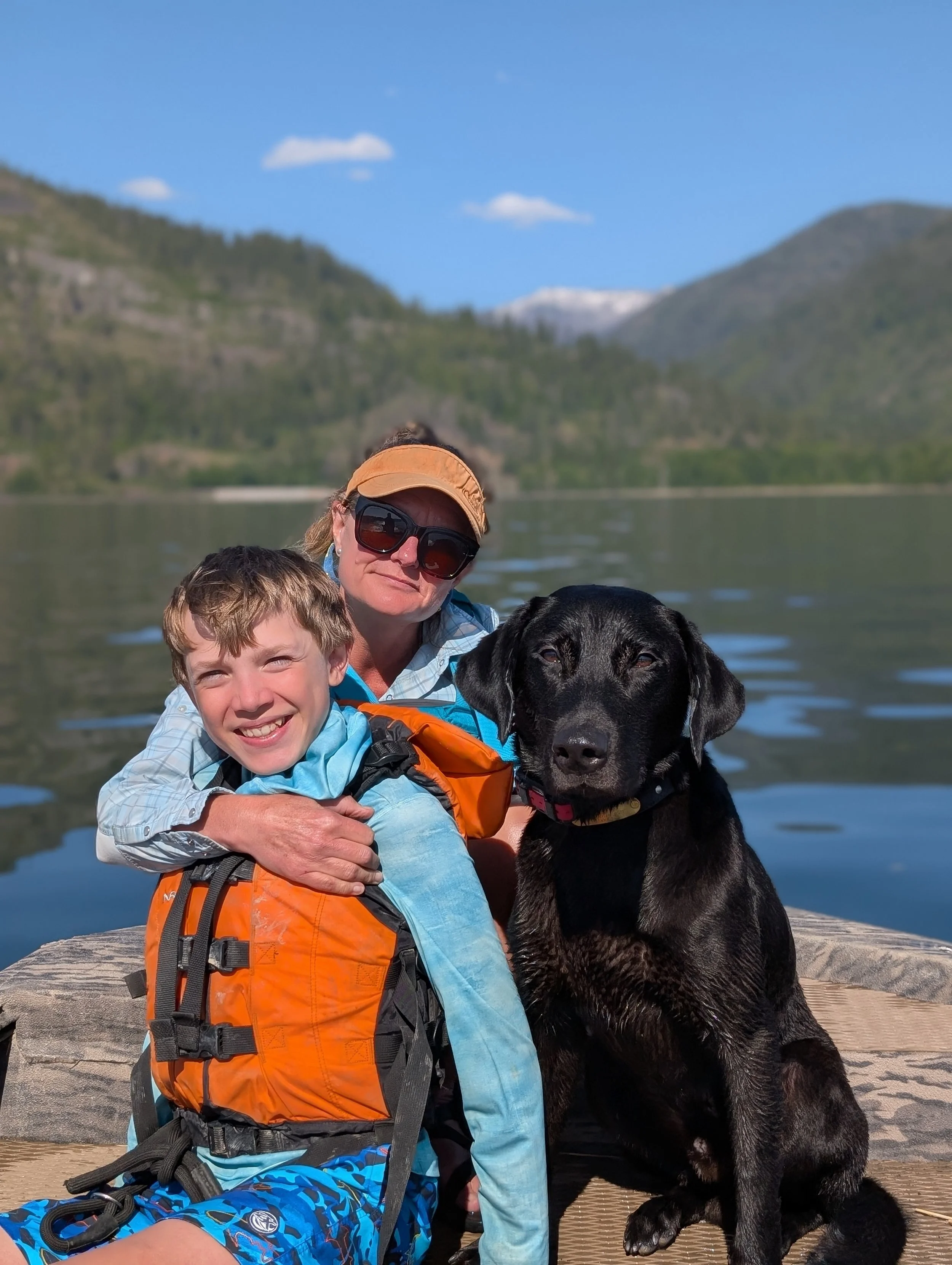 A woman, a boy, and a black dog sitting on a wooden dock by a lake with mountains in the background. The woman is wearing sunglasses and a tan cap, the boy is smiling and wearing a bright life jacket, and the dog is sitting calmly.