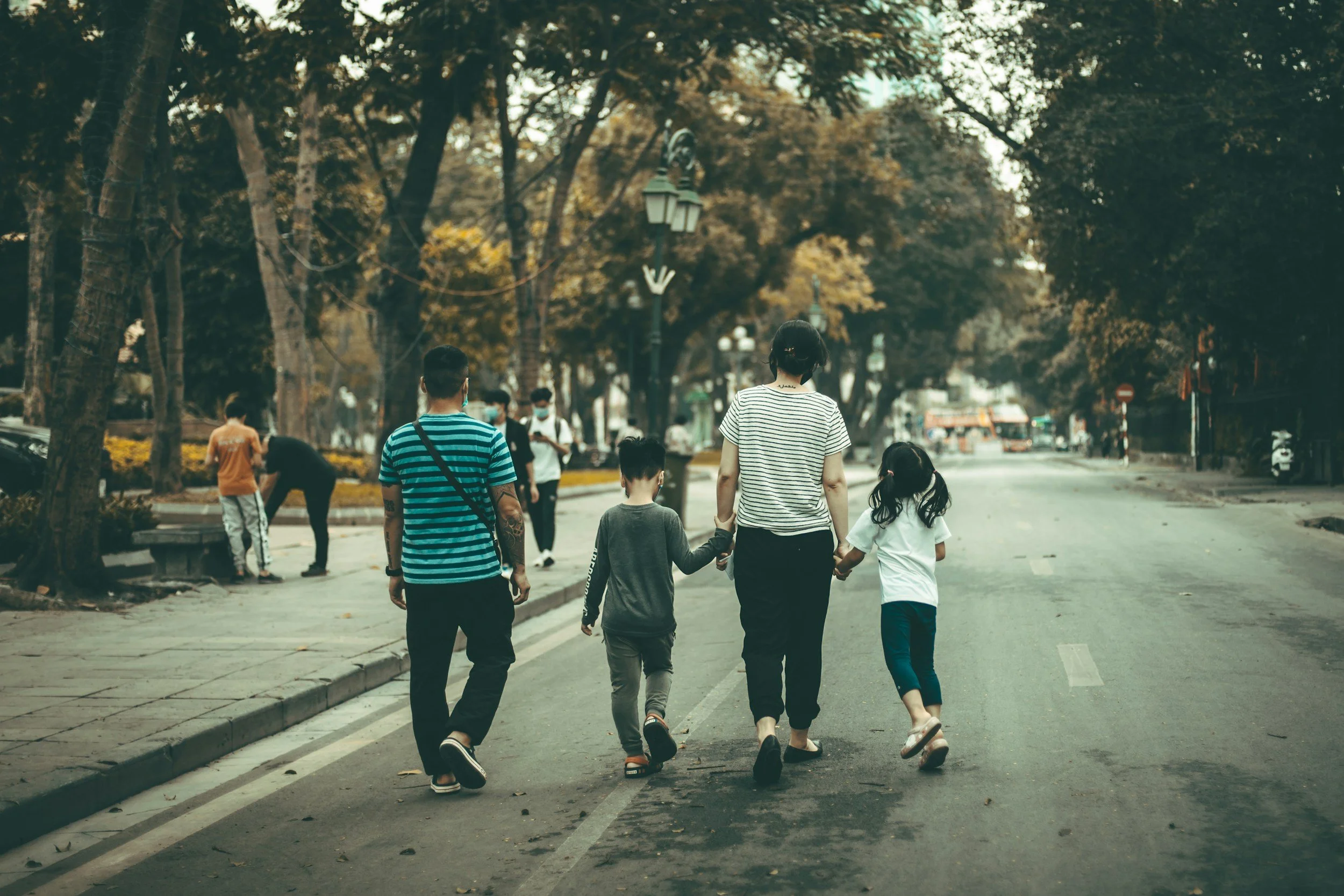 A family of four walking hand-in-hand down a city street, with other people in the background and trees lining the street.