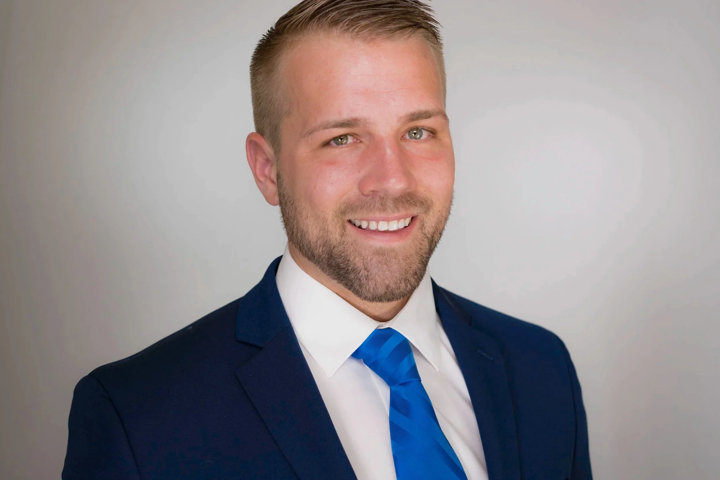 A professional headshot of a man with short, light brown hair, blue eyes, and a beard, wearing a dark blue suit, white shirt, and blue tie, smiling against a light gray background.