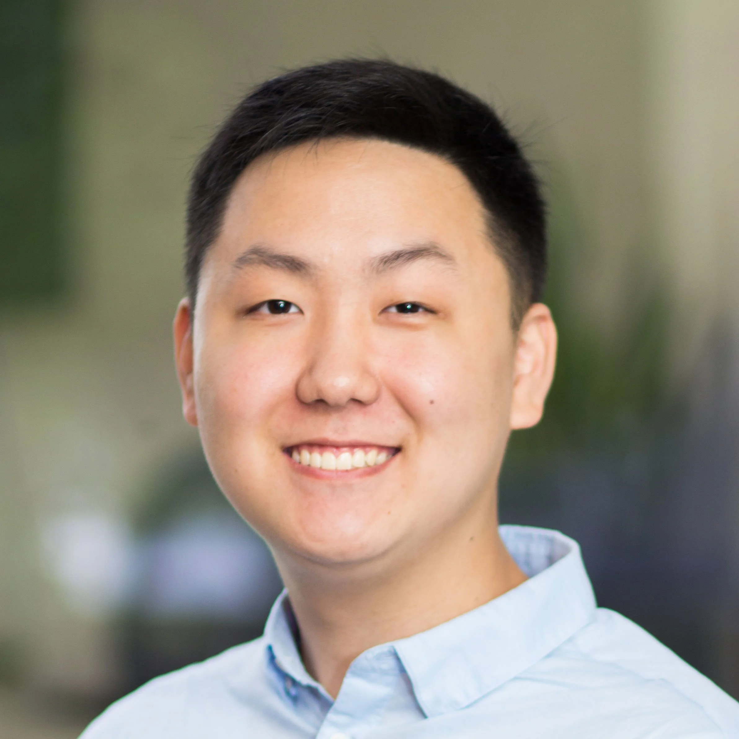 Headshot of a young Asian man with short black hair, smiling, wearing a light blue collared shirt, with a blurred outdoor background.